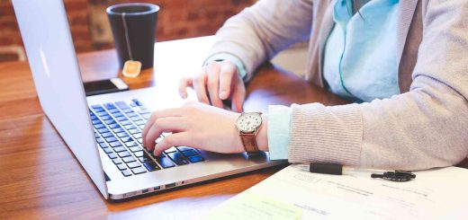 Na zdjęciu osoba siedząca przy biurku pracująca na laptopie, obok leży otwarta książka, po drugiej stronie stoi kubek